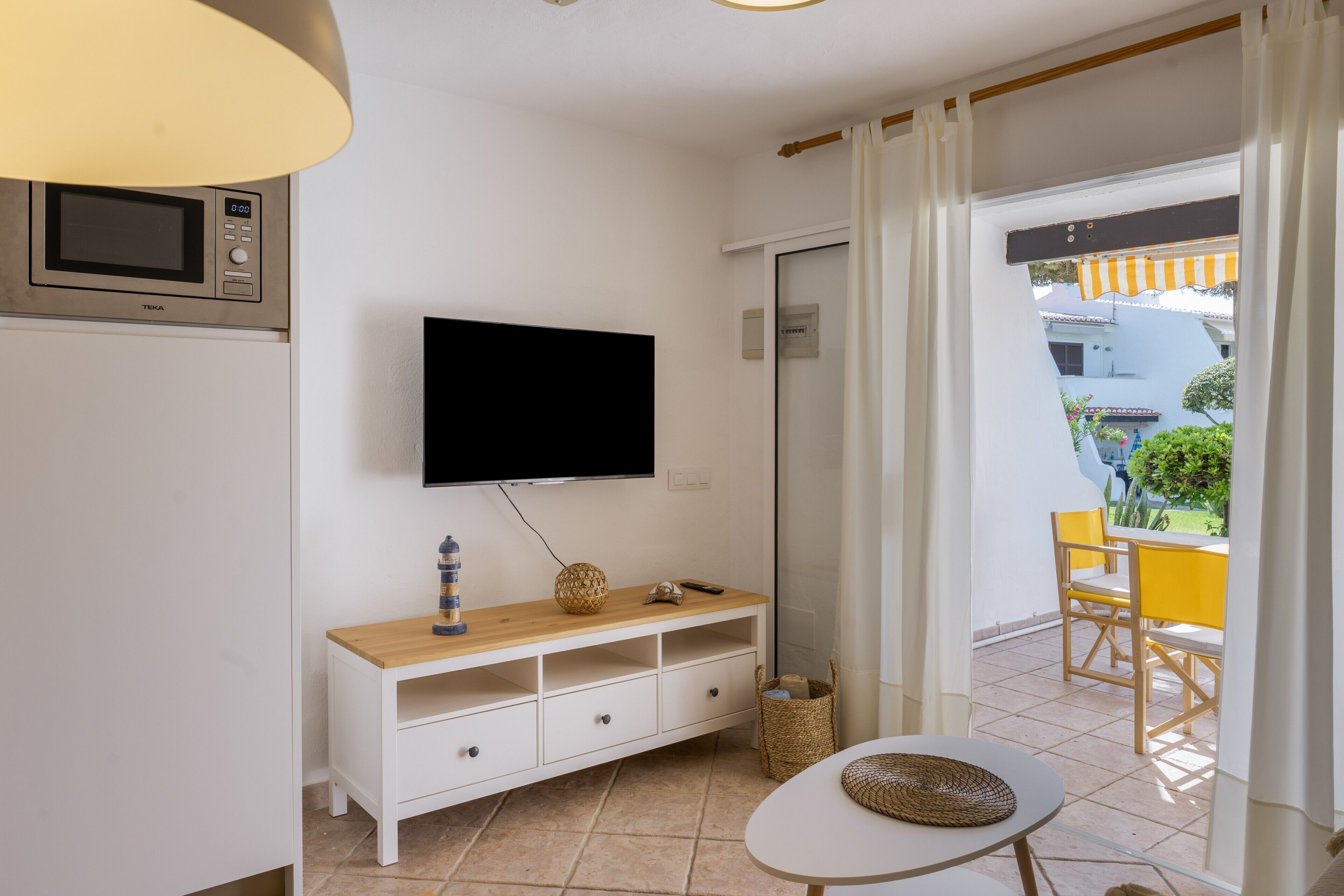 Una habitación con televisión montada en la pared, un aparador de madera con cajones, una mesita y una cesta de mimbre.