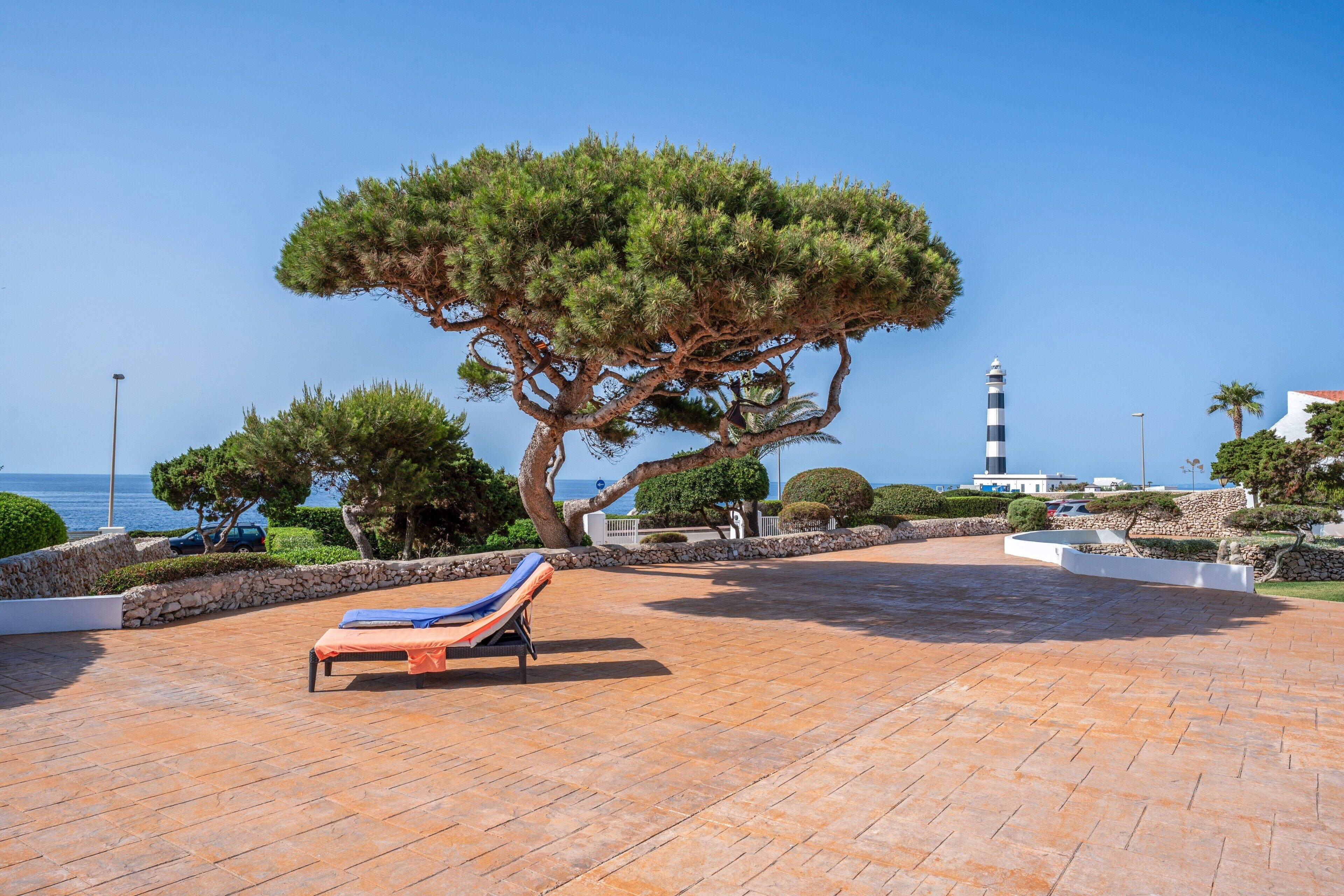 Un espace de détente extérieur, baigné de lumière, avec un grand arbre, un fauteuil et une vue sur la mer et un phare.