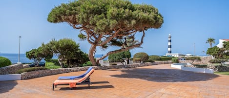 A sunlit outdoor lounge area with a large tree, a lounge chair, and a view of the sea and a lighthouse.
