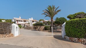 Una entrada cerrada a una zona residencial con edificios blancos, una palmera y arbustos podados.