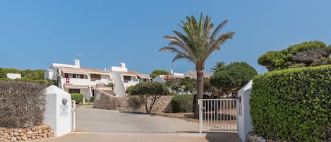 A gated entrance to a residential area with white buildings, a palm tree, and neatly trimmed bushes.