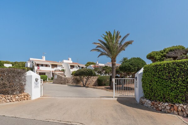A gated entrance to a residential area with white buildings, a palm tree, and neatly trimmed bushes.