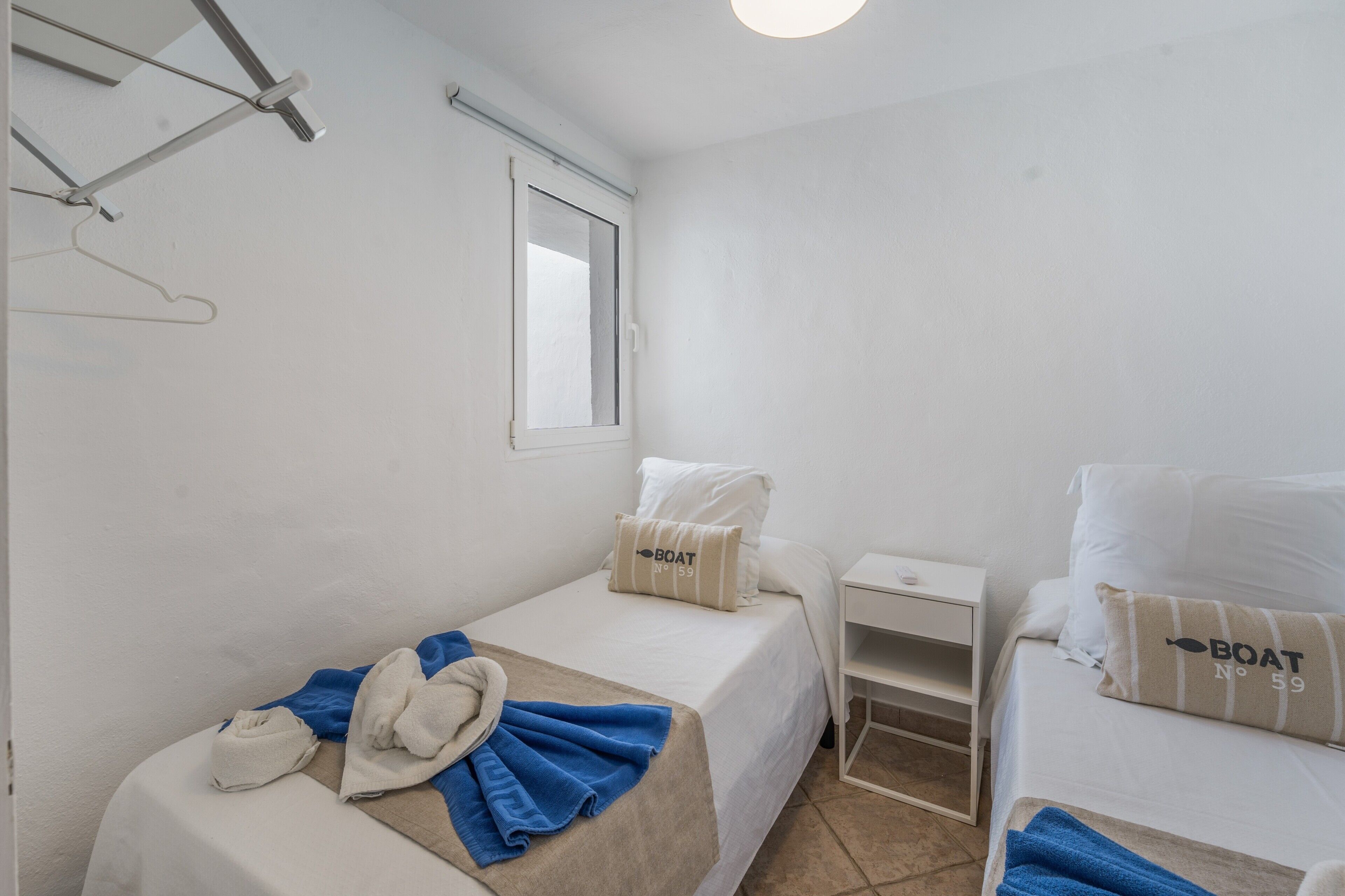 Habitación de hotel con dos camas, una mesita de noche pequeña, una ventana y un perchero para toallas en la pared.