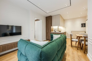A modern living room with a large flat-screen TV, a green upholstered sofa, a wooden dining table with chairs, and a kitchen area with wooden cabinets and a countertop. - Metropolitan Living 1BR City Apartment (Tirana)