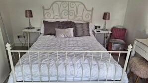 A bed with a white headboard, a bedside table with a lamp, a chair, and a small table with a lamp. - Bungalow - Sleeps 4 - Parking - Garden (Nottingham)