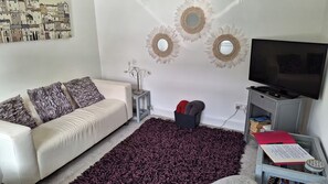 Un salon avec un canapé blanc, un tapis violet foncé, un téléviseur et des œuvres d’art murales.