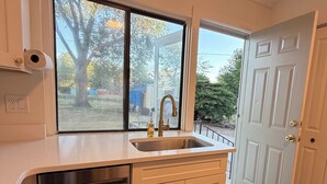 A kitchen with a stainless steel dishwasher, a sink, and a window with a view of trees and a blue tarp outside.