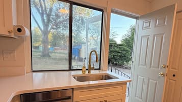 A kitchen with a stainless steel dishwasher, a sink, and a window with a view of trees and a blue tarp outside.