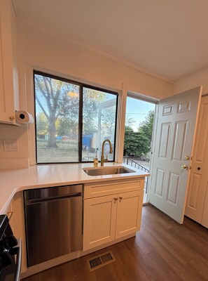 Una cocina con lavavajillas de acero inoxidable, un fregadero y una ventana que da a vistas de árboles y un toldo azul en el exterior.