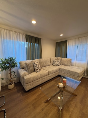 Una sala de estar con un sofá modular color crema, una mesa de centro de vidrio, una planta en maceta y una ventana con cortinas translúcidas.