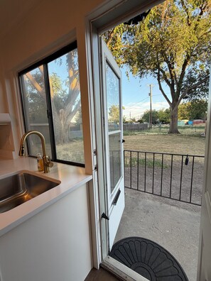 Una cocina con un lavamanos, una ventana y una puerta que da a un espacio exterior con un árbol y una valla.