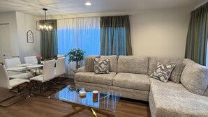 A living room with a sectional sofa, a glass coffee table, a dining area with a white table and chairs, and a ceiling with recessed lighting.