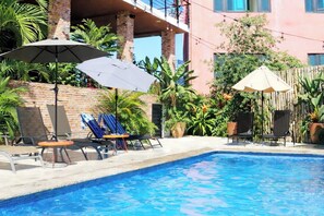 Une piscine entourée de chaises longues et de parasols, elle-même bordée d’une végétation luxuriante et d’un bâtiment en briques.
