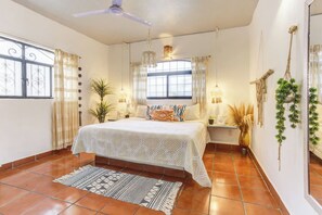 Une chambre à coucher avec un lit, un ventilateur de plafond, une fenêtre avec des rideaux, une table de chevet et une plante en pot.