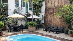 A pool area with lounge chairs, a table, and umbrellas, surrounded by potted plants and a wooden wall. - Pool Access • Walk to Beach • Kitchen • 
SAYULITA BOUTIQUE SUITES - 2 (Sayulita)