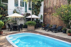 Ein Poolbereich mit Liegestühlen, einem Tisch und Sonnenschirmen, umgeben von Topfpflanzen und einer Holzverkleidung.