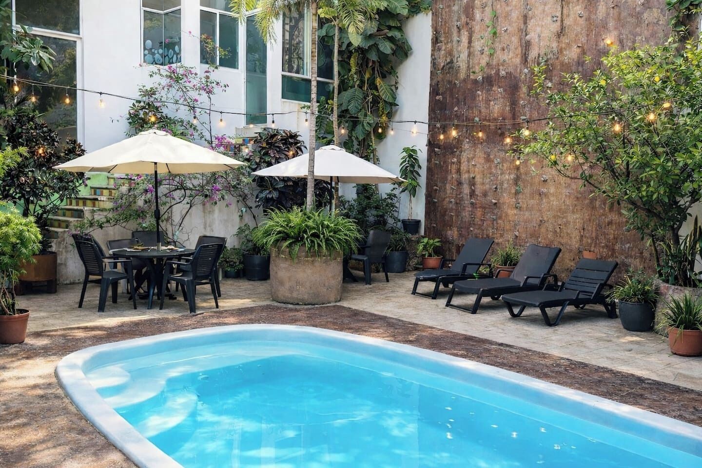 A pool area with lounge chairs, a table, and umbrellas, surrounded by potted plants and a wooden wall.