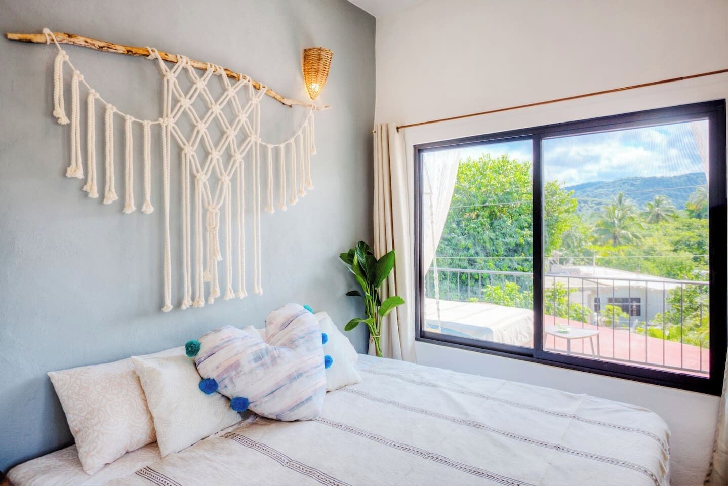 A bedroom with a bed, macrame wall decor, a potted plant, and a balcony with a view.