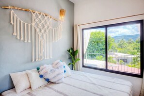 Une chambre avec un lit, des décorations murales en macramé, une plante en pot et un balcon avec vue.