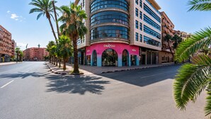 Un hotel moderno con una facciata curvilinea, palme che costeggiano la strada e negozi con insegne ben visibili.