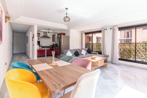Ein modernes Wohnzimmer mit Esstisch aus Holz, bunten Stühlen, einer Couch mit Kissen und einer Küche im Hintergrund.