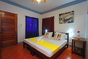 Habitación de hotel con una cama de madera, una colcha amarilla, un ventilador de techo, un cuadro en la pared y una puerta de madera.