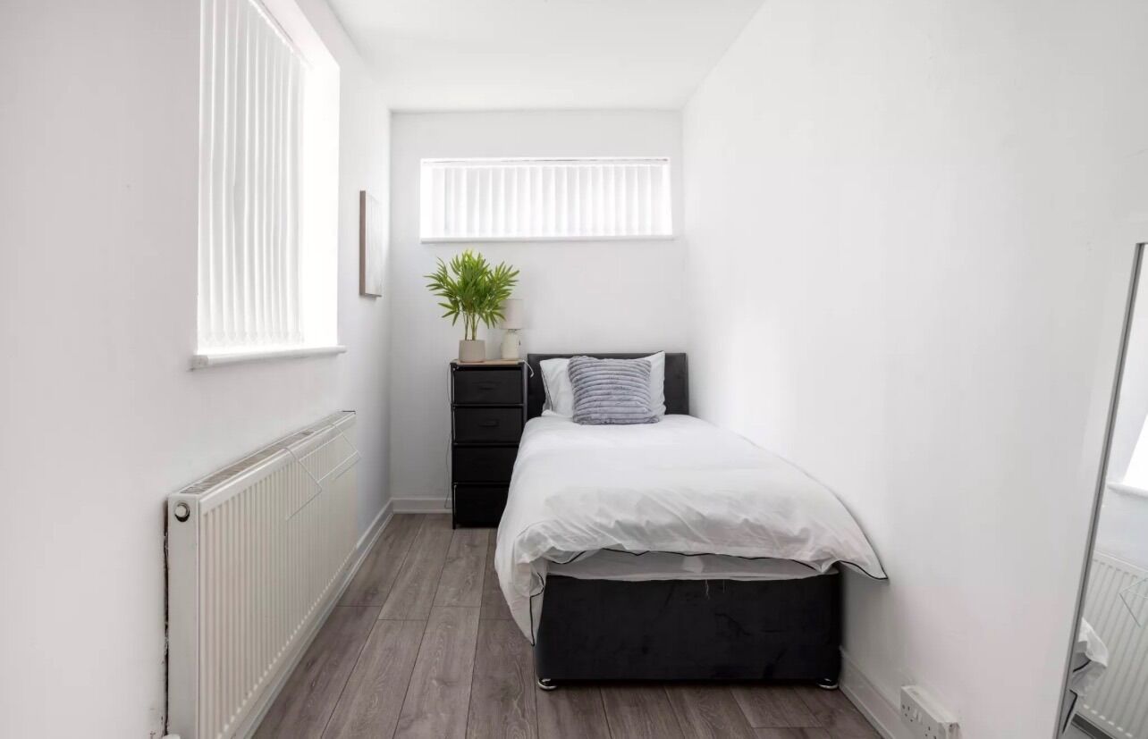 A narrow room with a bed, a radiator, a window with blinds, and a plant on a side table.