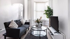 A modern living room with a sofa, a glass coffee table, a TV, and a potted plant. - House Sleeps 3 with Garden (Birkenhead)