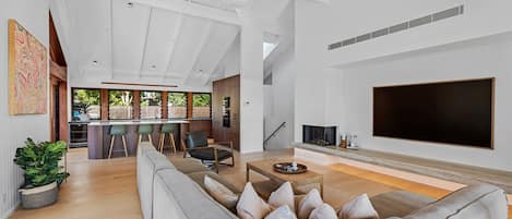 A modern living room with a large grey sofa, a flat-screen TV, and a kitchen area in the background.