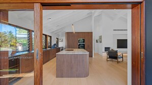 A modern kitchen with a central island, wooden cabinetry, and a view through a glass door. - 6 Karboora Drive - Luxury holiday home with pool (Point Lookout)