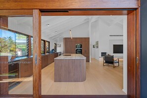 A modern kitchen with a central island, wooden cabinetry, and a view through a glass door. - 6 Karboora Drive - Luxury holiday home with pool (Point Lookout)
