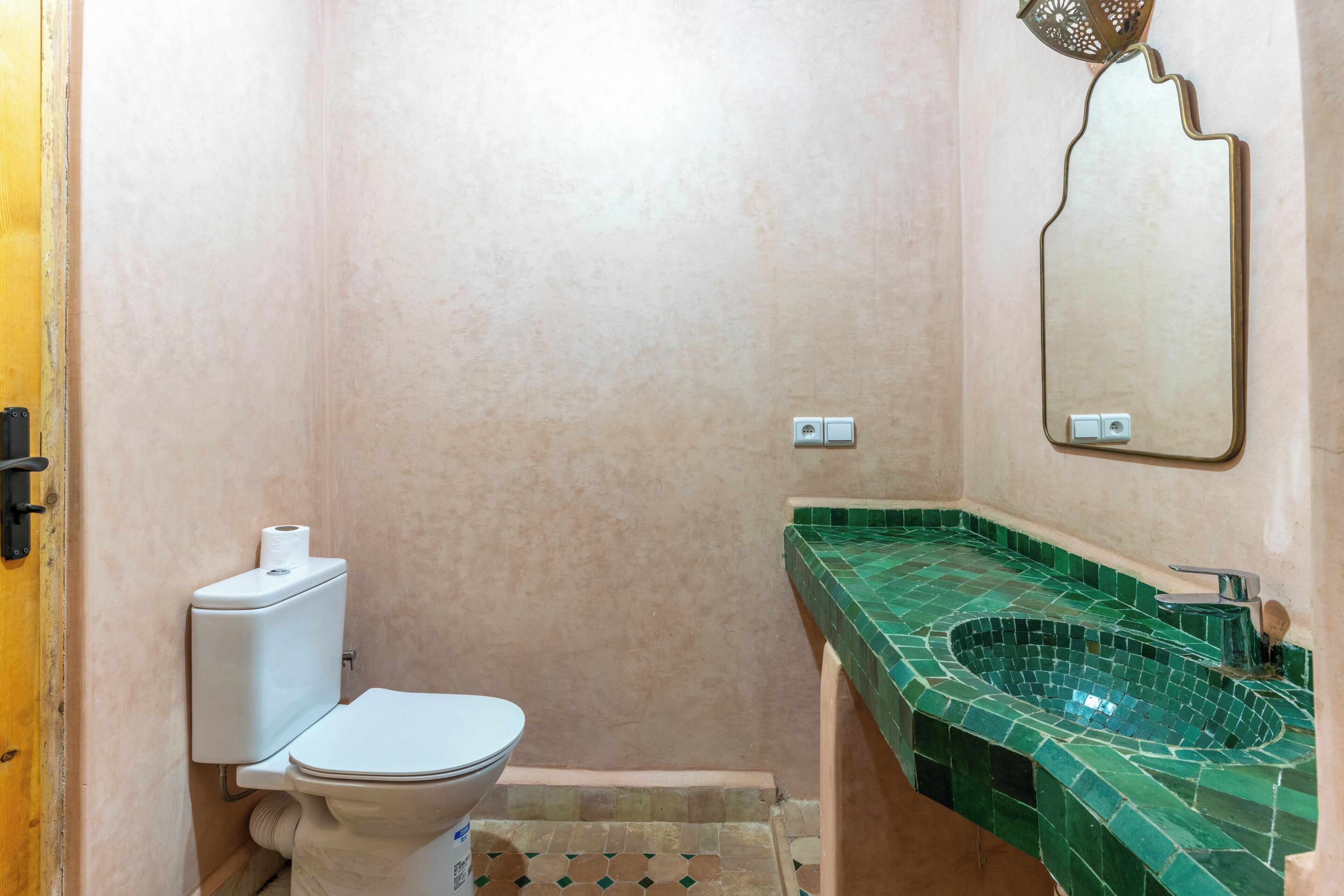 A bathroom with a toilet, green mosaic sink, and a wall-mounted mirror.
