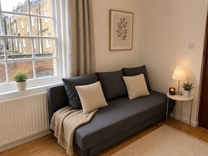 Una sala de estar moderna con un sofá gris, dos cojines, una manta color beige, una planta en marco, una mesita con una lámpara y una ventana con cortinas.