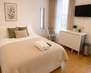 Une chambre d’hôtel bien rangée, avec un lit, un bureau, une chaise, une télévision et une poubelle.