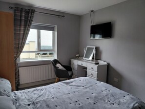 A bedroom with a bed, a desk, a chair, a radiator, a window with curtains, and a TV mounted on the wall.