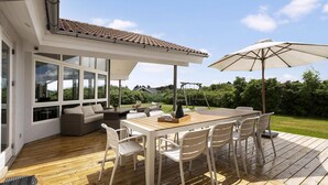 Een overdekt terras buiten met een houten tafel en stoelen, een schommel en uitzicht op de omliggende groen.