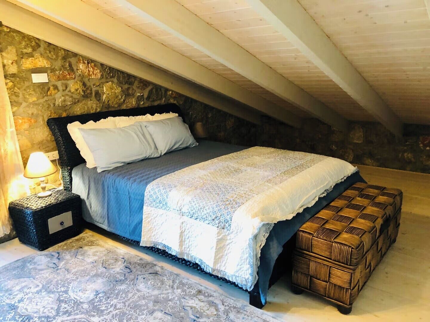 A bedroom with a stone wall, a bed with a white and blue bedspread, a bedside lamp, and a wicker bench.