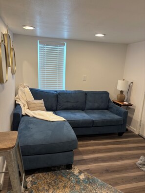 Ein Wohnzimmer mit einem blauen Modulsofa, einem kleinen Beistelltisch mit Lampe, Holzboden und einem Fenster mit Jalousien.