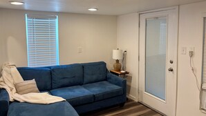 A room with a blue sofa, a white door, a window with blinds, and a lamp on a side table. - Cozy lower-level 1 bedroom studio in NOLA with Private Covered Outdoor Patio (New Orleans)