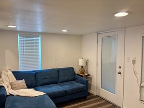 A room with a blue sofa, a white door, a window with blinds, and a lamp on a side table. - Cozy lower-level 1 bedroom studio in NOLA with Private Covered Outdoor Patio (New Orleans)