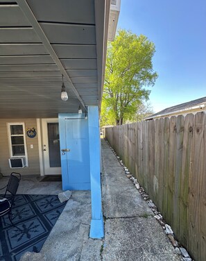 Ein überdachter Patio-Bereich mit einer blauen Tür, einem Holzzaun und wolkenlosem Himmel.