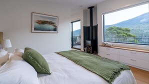A bedroom with a bed, a green throw, a fireplace, a window with a mountain view, and a painting on the wall.
