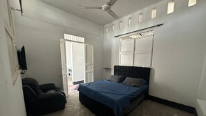 Une chambre à coucher avec un lit, un ventilateur de plafond, une fenêtre avec des stores et une porte donnant sur une autre pièce.