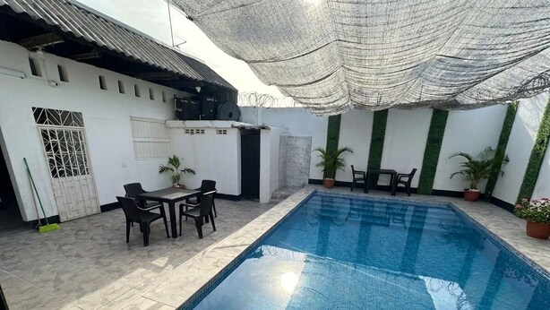 Une piscine avec un revêtement de sol carrelé, un espace détente couvert avec des chaises et des plantes en pot.