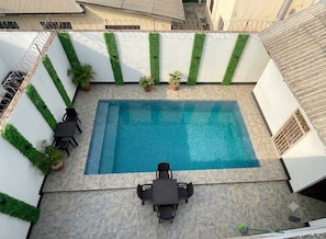 A rooftop swimming pool with a tiled deck, surrounded by white walls with green vertical garden accents, and a small table with chairs.