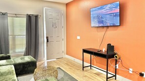 Une pièce avec un téléviseur fixé au mur, une porte, une fenêtre avec des rideaux, un canapé et une petite table avec un haut-parleur.