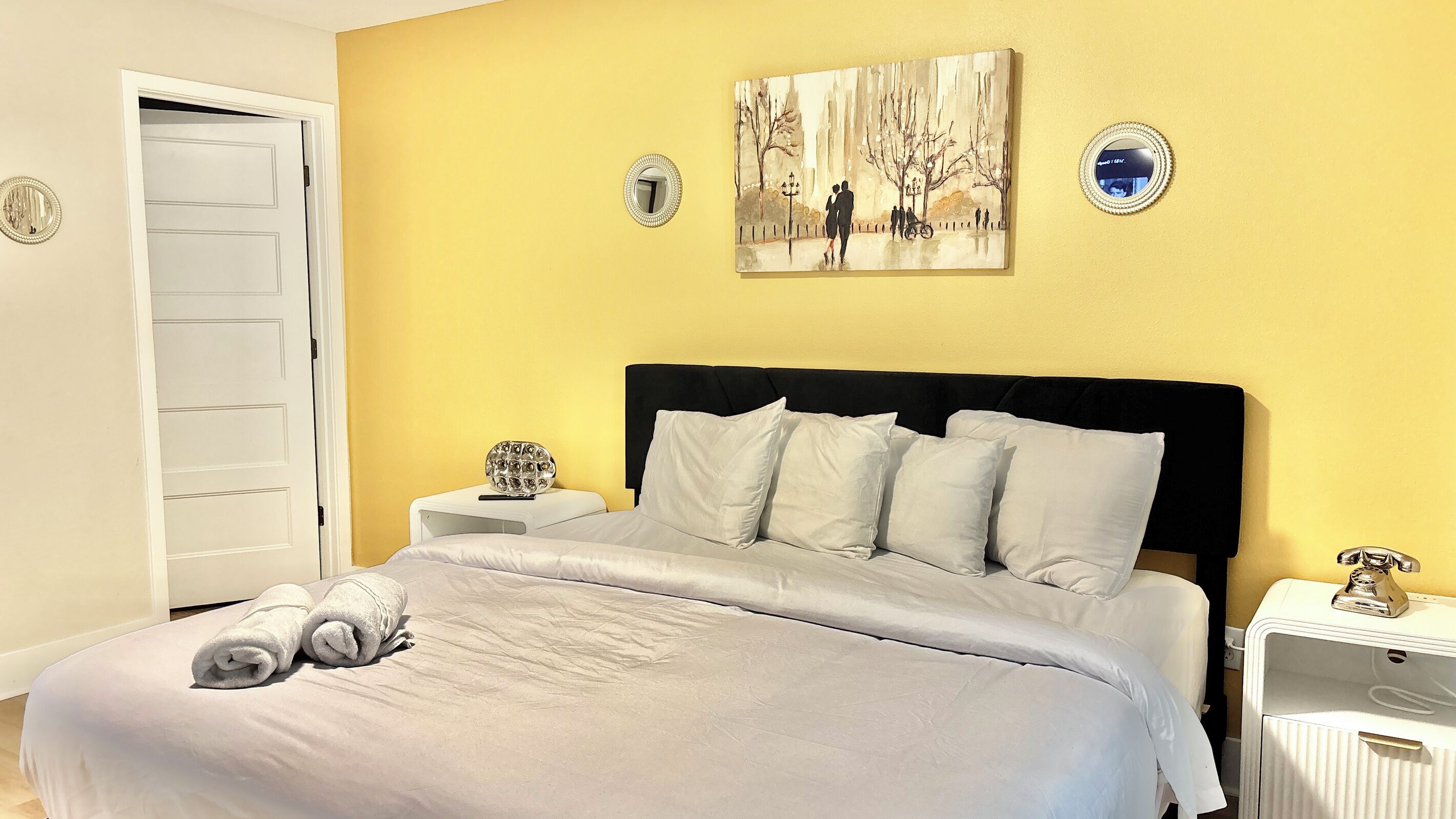 A hotel room with a bed, a nightstand, a bedside table, a mirror, and a painting on the wall.