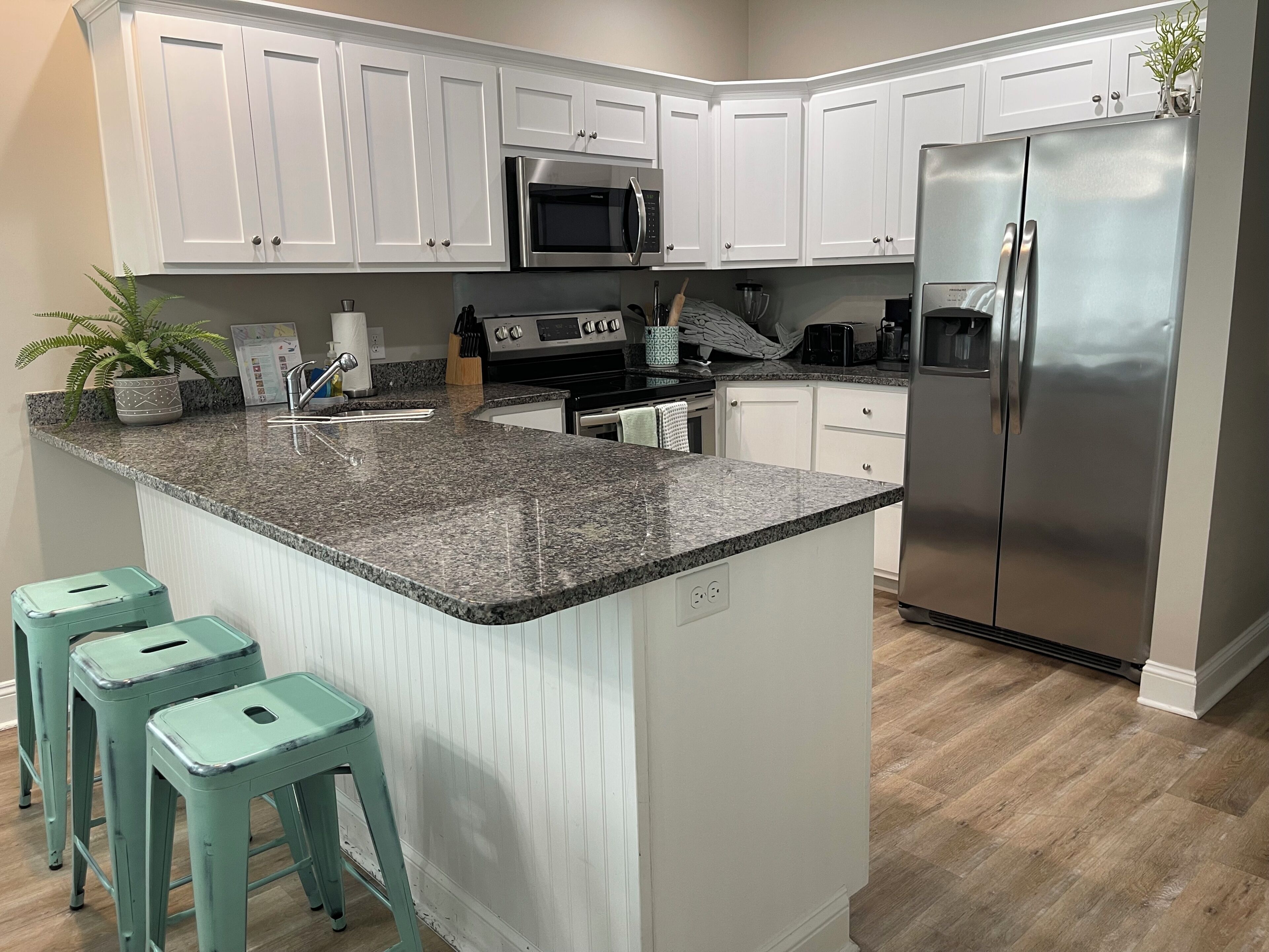 A kitchen with white cabinets, a stainless steel refrigerator, a granite countertop, and teal bar stools.