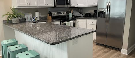 A kitchen with white cabinets, a stainless steel refrigerator, a granite countertop, and teal bar stools.