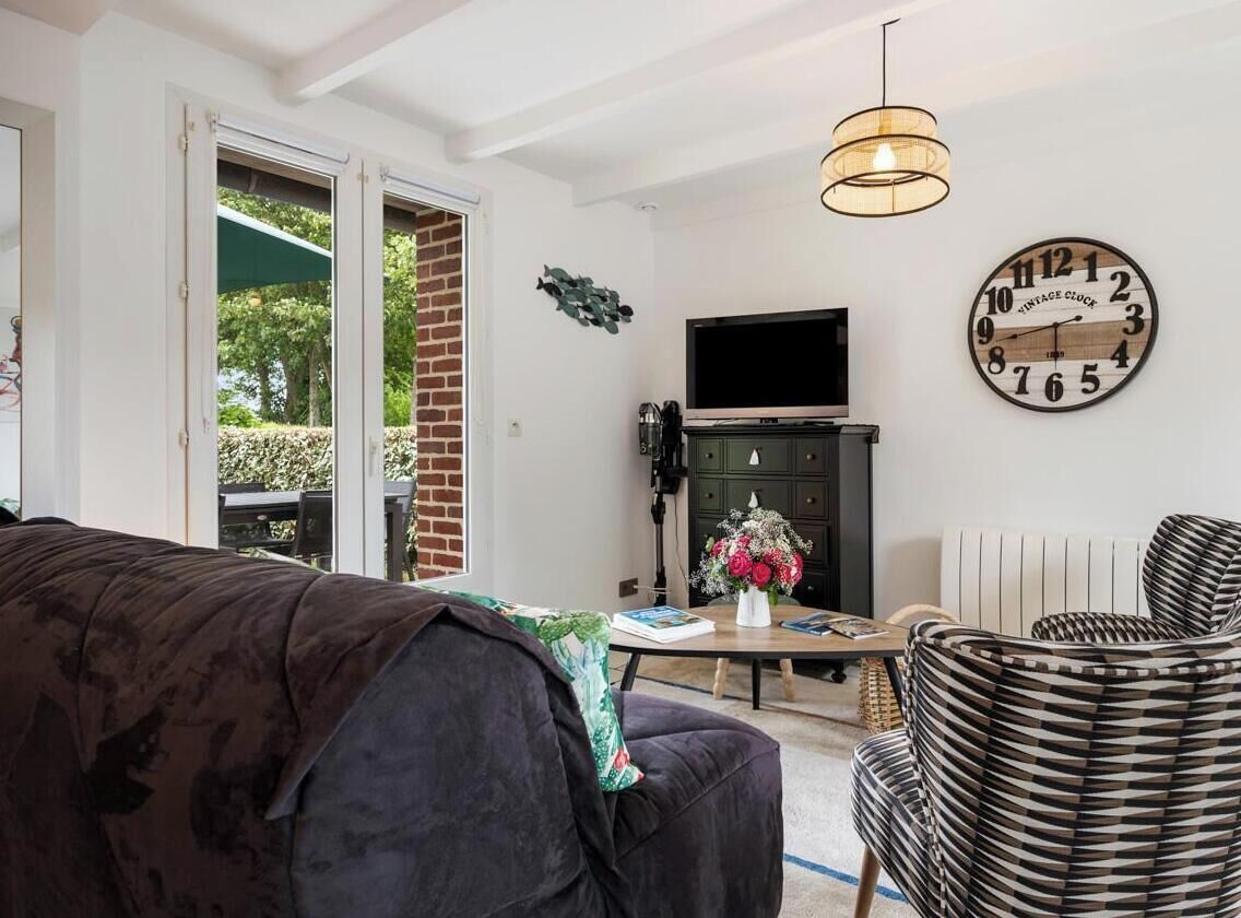 Un salon avec un canapé, un téléviseur, une horloge et une table sur laquelle est posé un vase de fleurs.